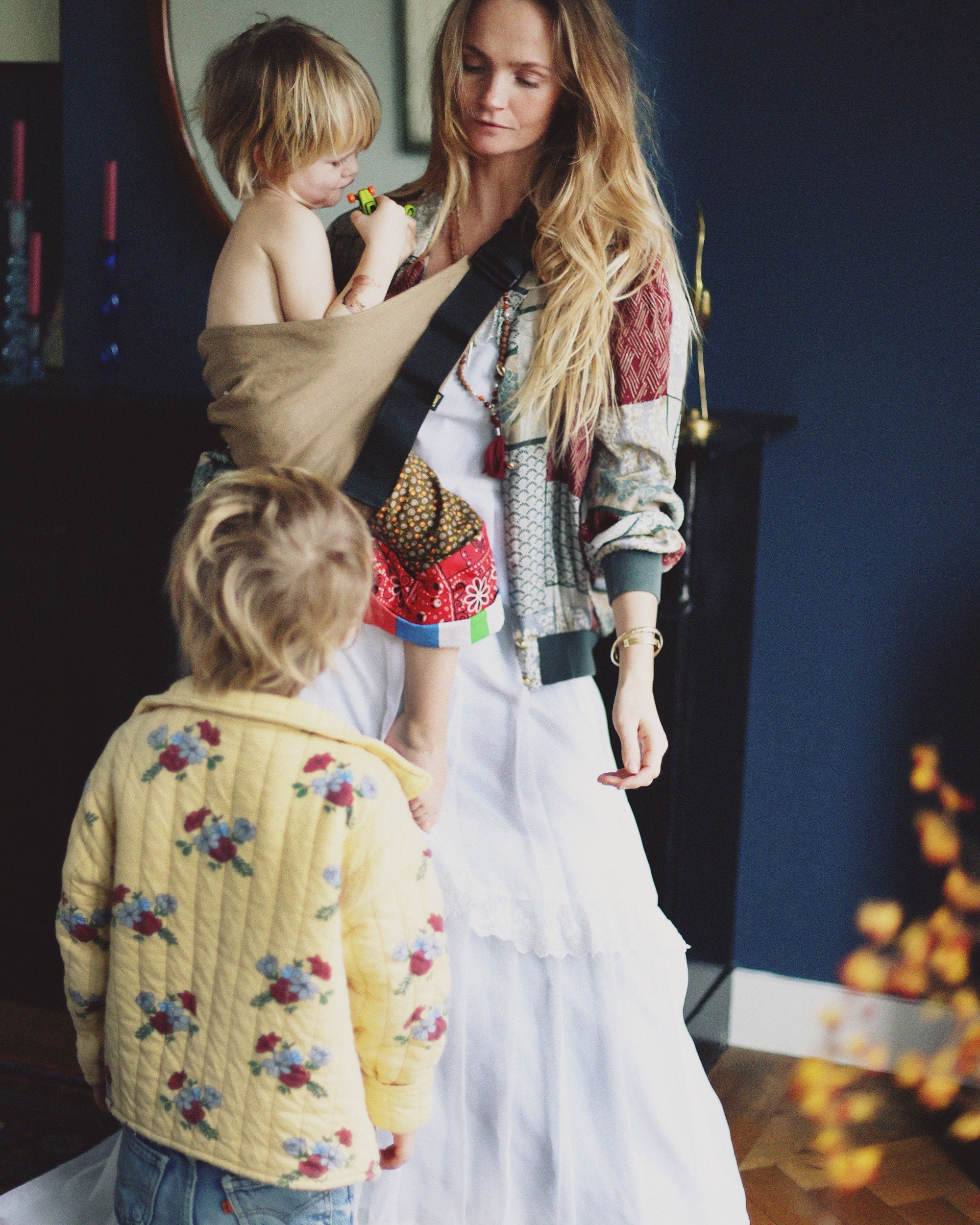 A mother wearing the Wildride Camel toddler carrier at home, holding a child while another stands nearby.