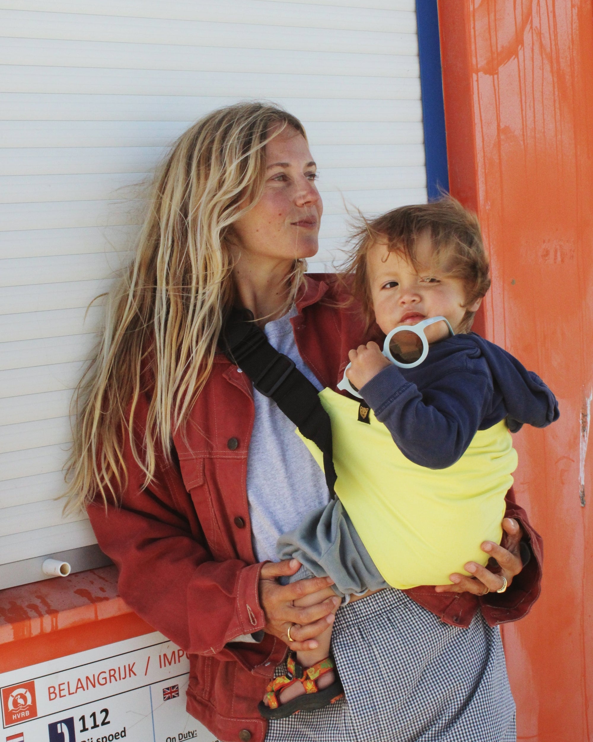 A mother wears a Wildride toddler carrier in Lemon, holding her child close in a hip carrier. The toddler, in a navy hoodie, looks at the camera while gripping a pacifier. The best baby carrier is secured with a black strap, offering hands-free comfort.