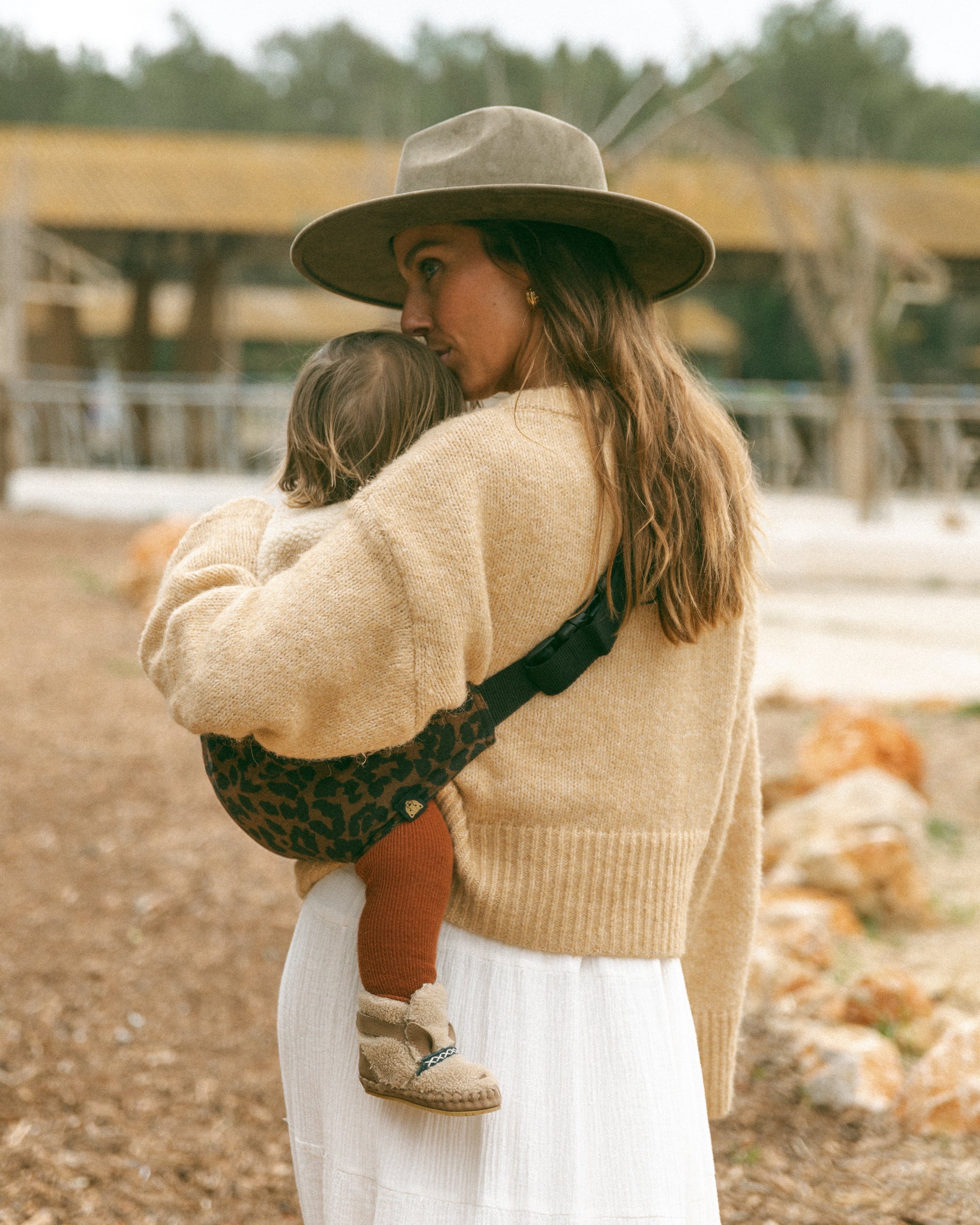 Woman in a beige sweater and hat carrying a toddler on her hip in a Wildride brown leopard print toddler carrier — ergonomic hip carrier for everyday use
