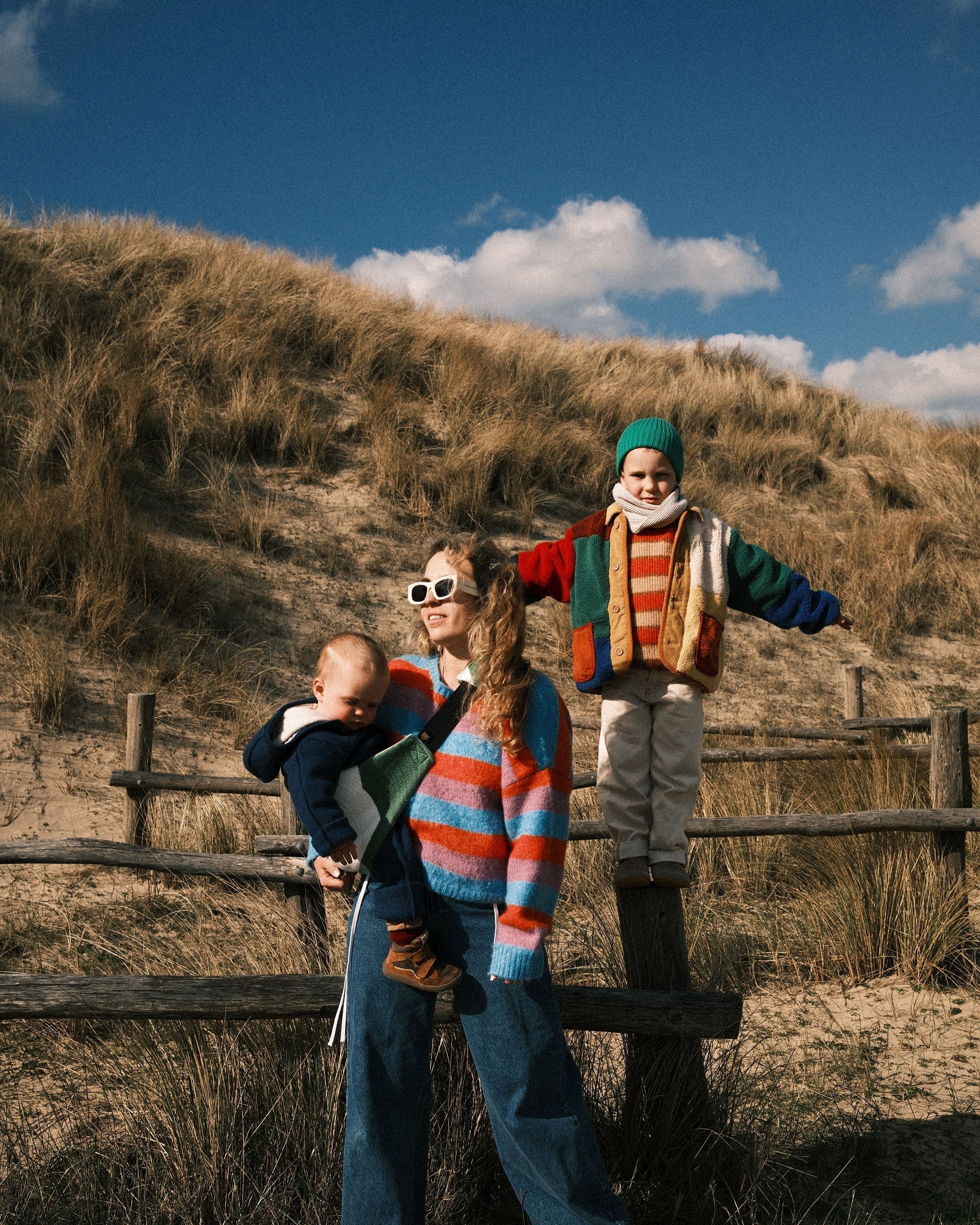 Mother standing in the dunes with her baby in a Wildride baby carrier, while her older child balances on a wooden fence nearby. A calm family moment with their toddler carrier.