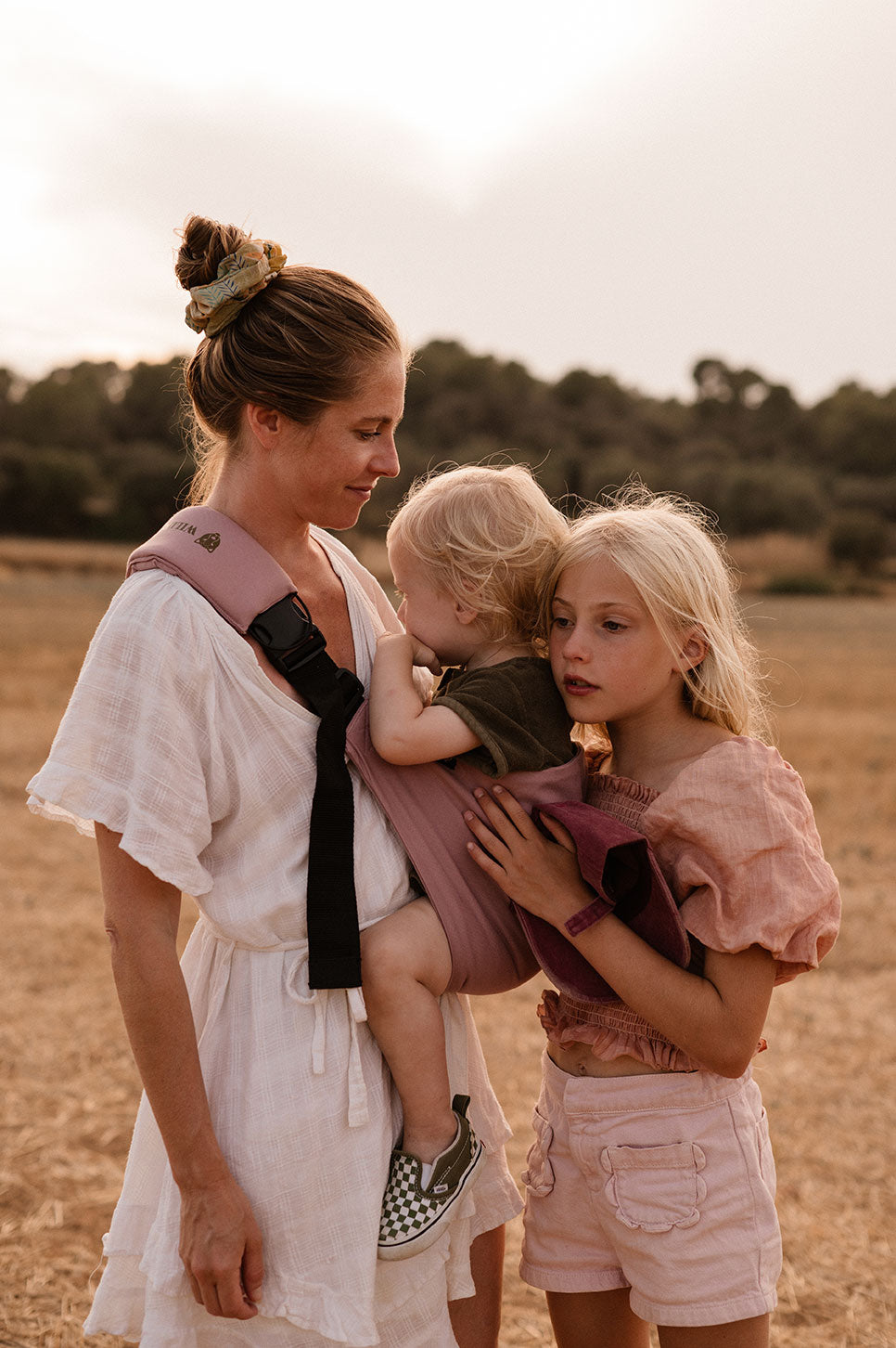 A woman stands in a golden field carrying her child in a Wildride toddler carrier, one of the best baby carrier options for outdoor adventures, while an older child stands close beside them.