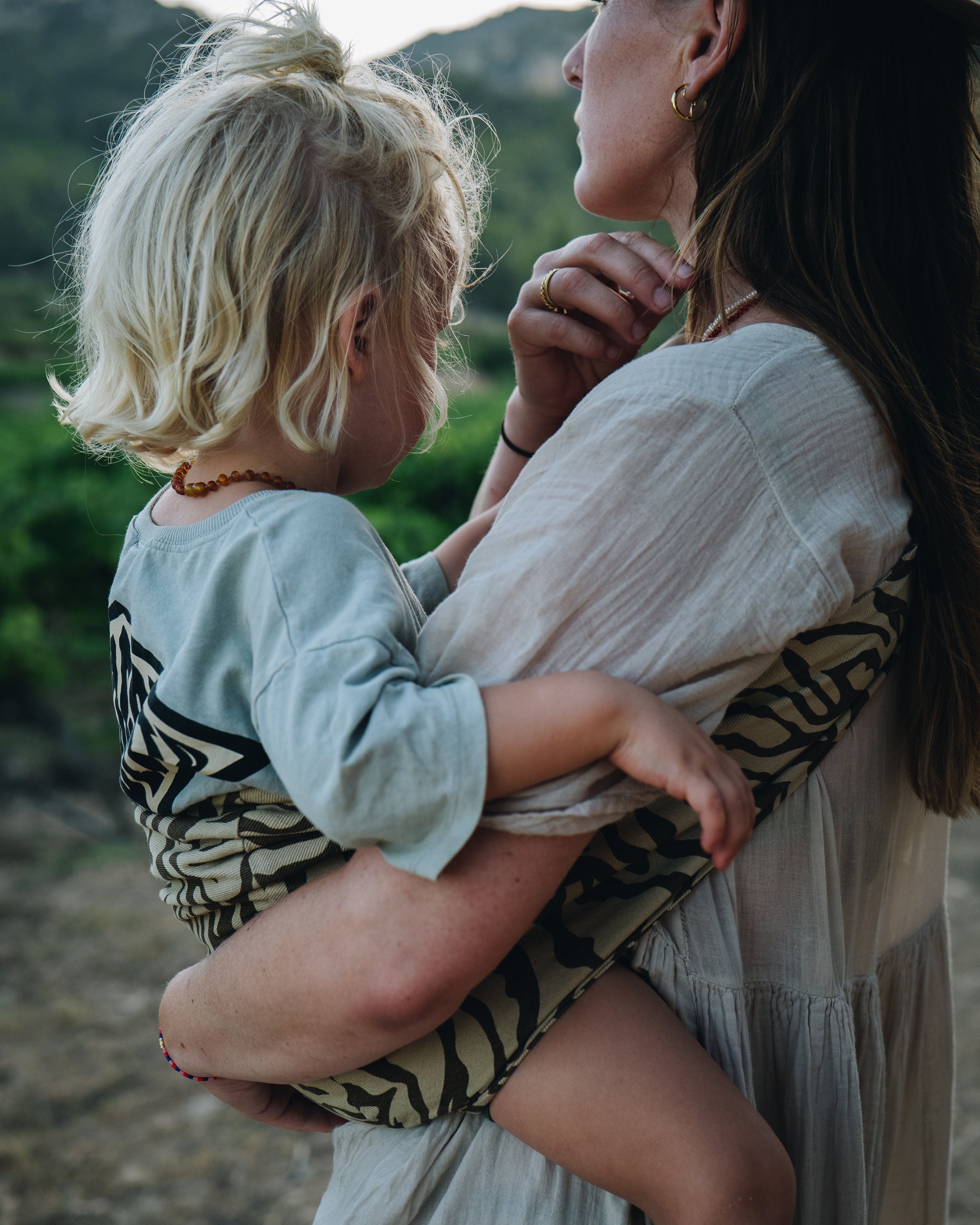 A close moment between mother and child, captured outdoors. The child is carried in a zebra-print Wildride hip carrier as they share a quiet look together.