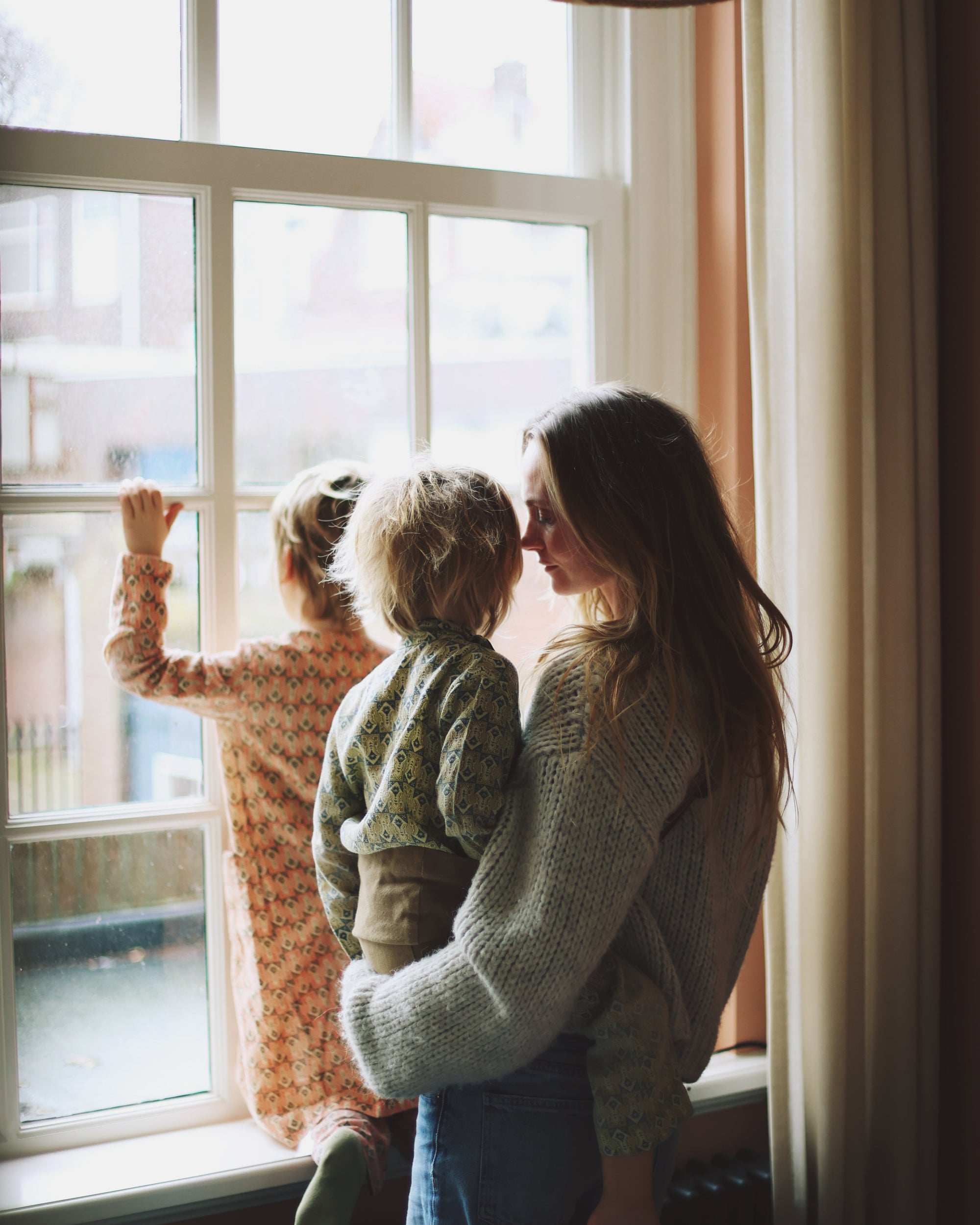 A mother holds her toddler in a Wildride toddler carrier while standing by a window, with another child looking outside. Natural light fills the room.