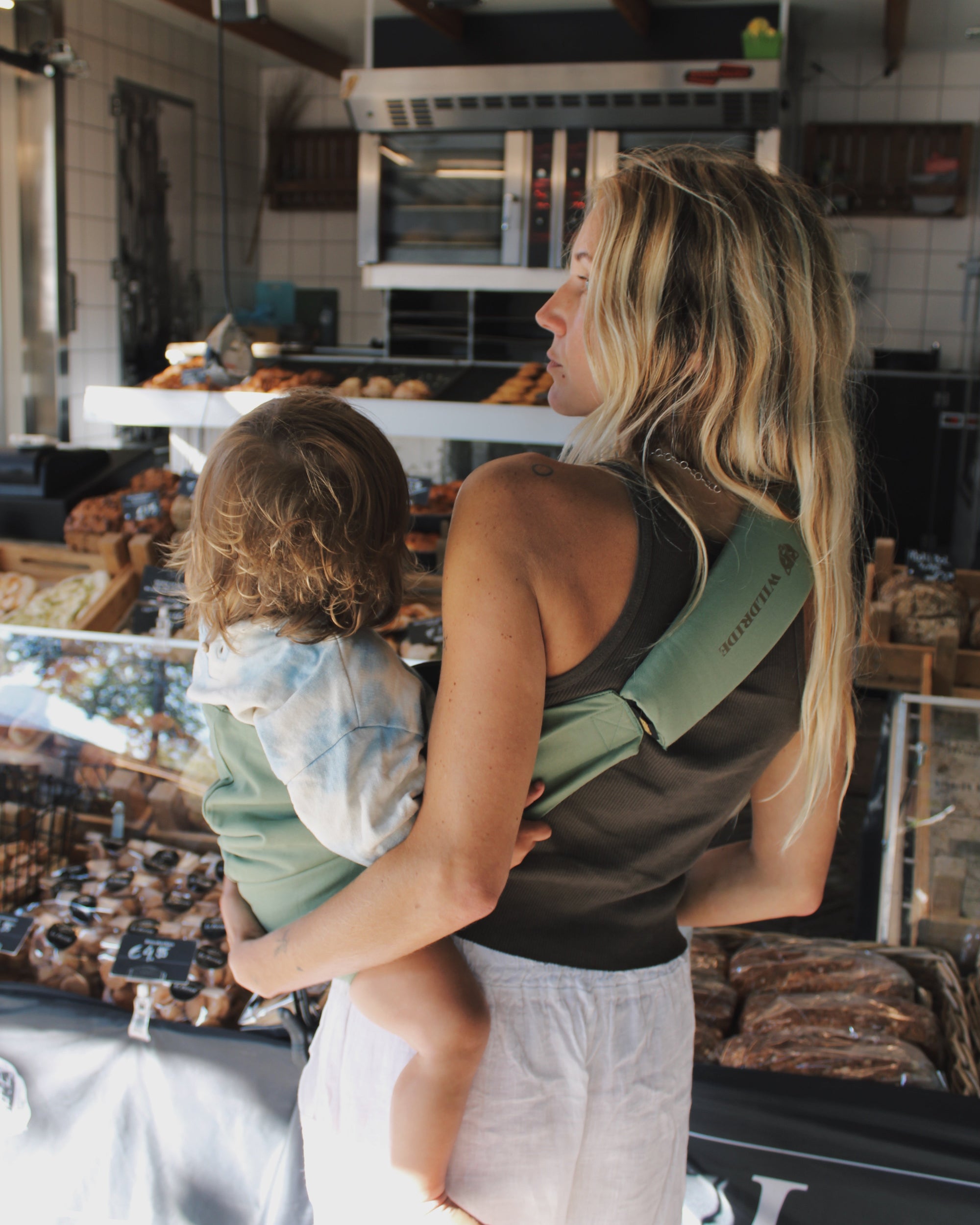 Mother carrying child in Wildride Fern carrier at a bakery counter
