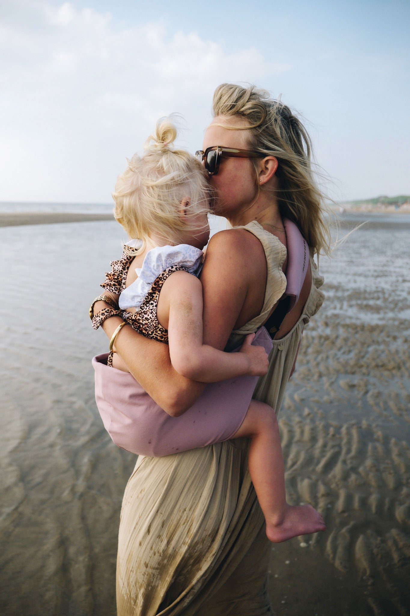 Mother carrying her child in a Wildride Toddler Carrier Vintage Rose, a lightweight ergonomic hip carrier, standing on the beach at low tide.